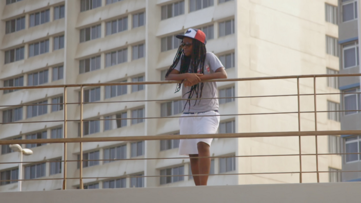 Suthu Magiwane is standing at a railing. Her long dark hair comes out from under her red baseball cap. She is wearing sunglasses. A high-rise building stands in the background.