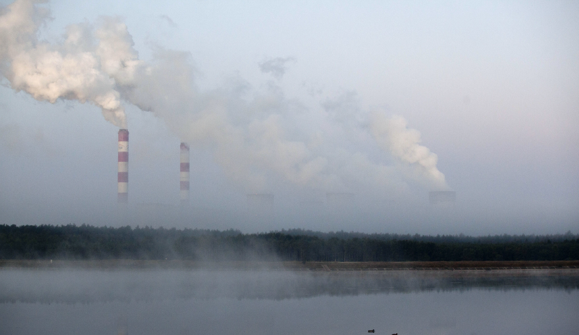 High plumes of smoke come out of two tall red and white striped chimneys and three wide chimneys. There is a forest in front of the chimneys and a body of water in front of them.