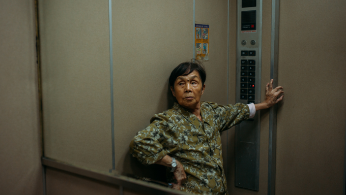 Frau Tu ist eine Dame mittleren Alters. Sie lehnt an der Wand eines Fahrstuhls und stützt sich mit einer Hand ab.