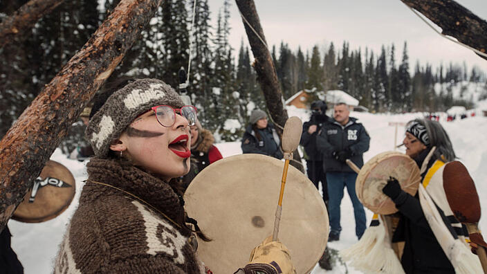 Three women stand in a snowy landscape surrounded by trees. The women are singing and drumming. Three other people stand at the edge and watch the women.