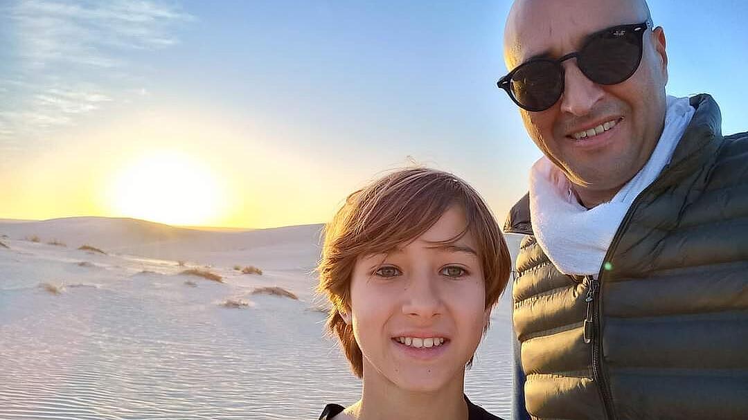 Ein Porträt eines Mannes mit dunkler Sonnenbrille und eines Jungen. Sie stehen in einer Wüstenlandschaft. Die Sonne geht am Horizont unter.