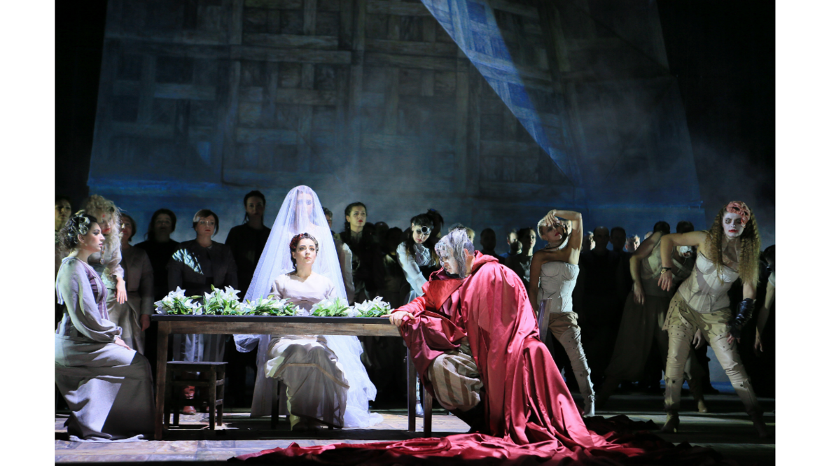 Auf einer Bühne sitzen zwei Frauen und ein Mann in antiken Kleidern an einem Tisch, hinter ihnen stehen Chormitglieder und Tänzer. Alle tragen Kostüme und sind geschminkt