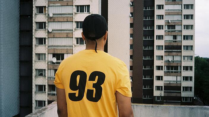 Mann mit gelbem T-shirt und Basecap steht auf einem Balkon, sein Blick ist auf die gegenüberliegende Hochhauswand gerichtet, auf seinem Rücken steht die Nummer 93