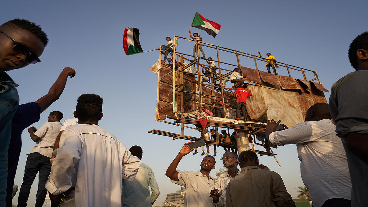 Mehrere Männer stehen im Freien herum. Mehrere Menschen klettern auf ein improvisiertes altes Gerüst, das in den Himmel ragt. Oben schwenken zwei junge Männer sudanesische Flaggen
