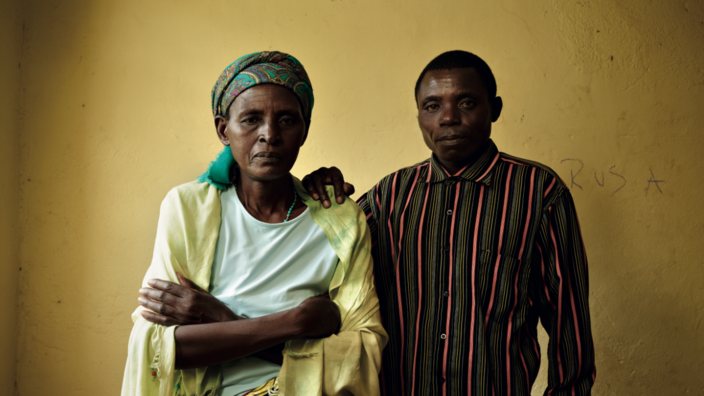 An old woman stands next to a middle-aged man. Both are from Rwanda. She has her arms folded in front of her chest. He has put his right hand on her left shoulder. He looks seriously into the camera. Her look is serious and bitter. She wears a brightly patterned scarf on her head, a brightly patterned skirt, a mini-green shirt and a bright yellow shoulder scarf. He wears a lengthwise striped shirt in brown, red and mustard yellow. The two are standing in front of a mustard yellow wall.