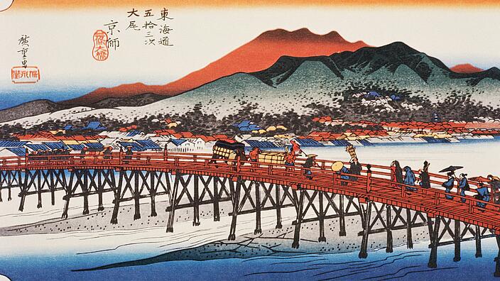 Illustration shows a wooden bridge over a river. Several people are crossing the river with luggage and carts. In the background there is a city of low houses and two mountains