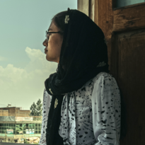 A young woman with a black headscarf and glasses sits sideways in the frame of a wooden entrance door. Through the door you can see several flat buildings and a few trees. The woman is looking towards the horizon. In her lap is an open book. She wears a white dress with a black pattern. 