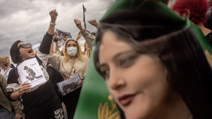 In the background, several women in a crowd raise their fists in the air and shout. They hold signs in their hands. In the foreground is a flag with the portrait of the Iranian Kurdish woman Mahsa Amini