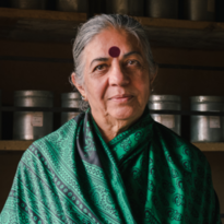 Vandana Shiva wears a green Sari. She is standing in front of a shelf with many tins and looks calmly into the camera 