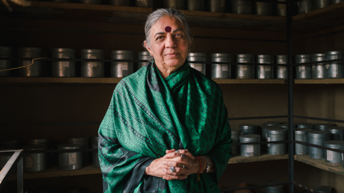 Vandana Shiva wears a green Sari. She is standing in front of a shelf with many tins and looks calmly into the camera 