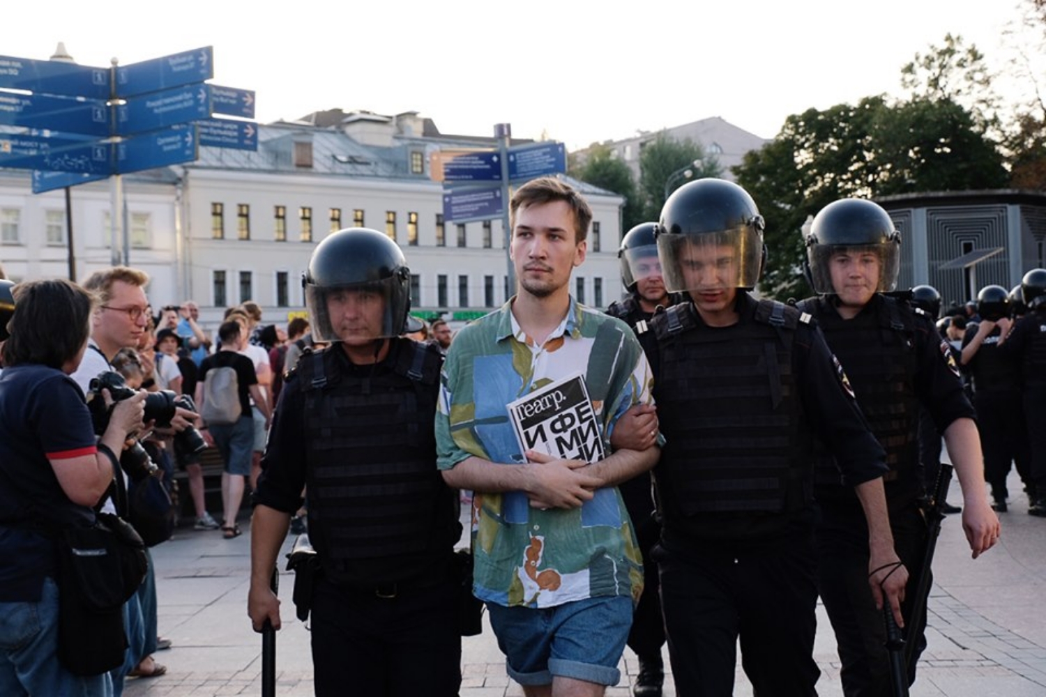 Ein junger Mann mit kurzen blonden Haaren und buntem Hemd wird von behelmten Polizisten in schwarzer Uniform abgeführt.