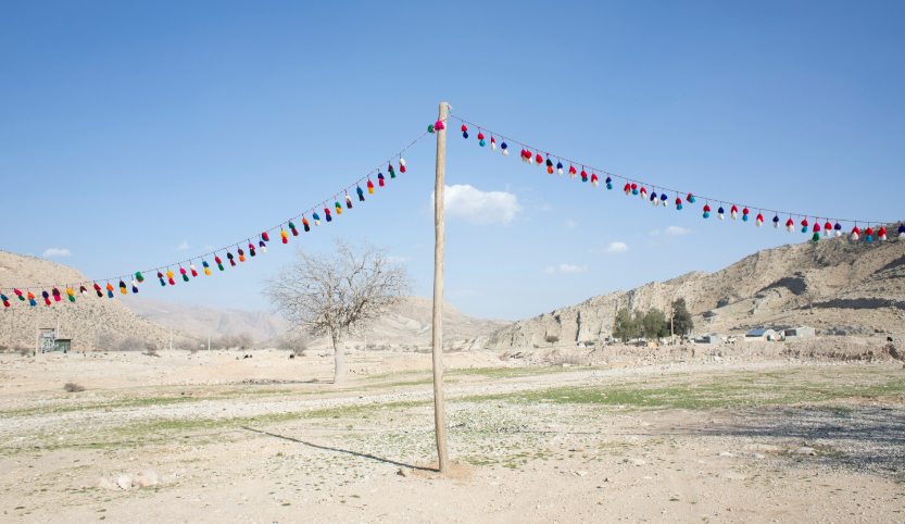 A colorful tassel garland hangs on a wooden pole in the middle of a barren landscape. In the background are mountains.