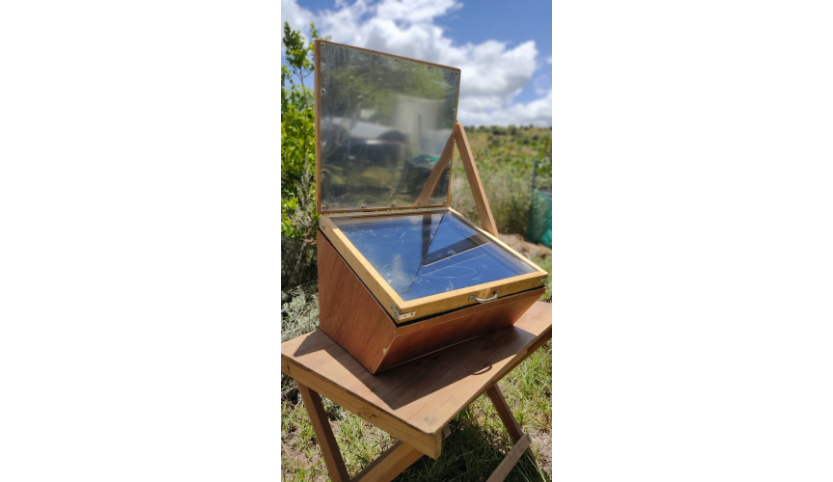 On a wooden table is a square wooden box with an open lid. The lid is covered with aluminum on the inside. Inside the box is another lid with window glazing. Through the window you can see two cooking pots.  